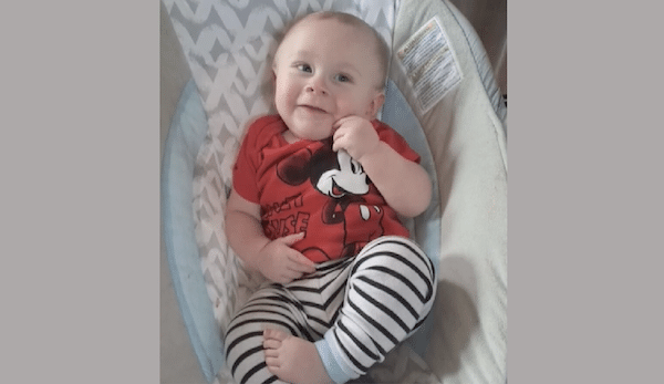 Baby boy in a red shirt featuring Mickey Mouse grins while lying back on a soft, light-colored baby seat.