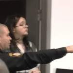 Defendant in gray-and-white striped attire is escorted by an officer during a court hearing, with a blurred background.