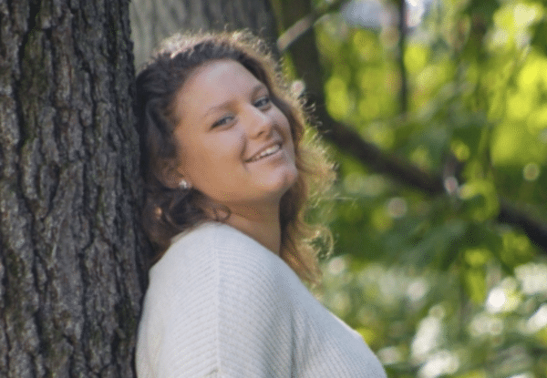 Close-up of a woman leaning on a tree trunk, smiling gently, surrounded by sunlit foliage.