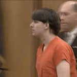 Side view of a person dressed in orange jail attire standing next to another individual in formal clothing, with a courtroom setting behind them.