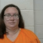 Woman in an orange correctional outfit and spectacles stands in front of a neutral wall for a mugshot image.