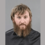Man with a thick, long beard and unkempt brown hair, posed for an official identification photo with a neutral gray background.