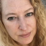Portrait of a woman with light skin and shoulder-length wavy blond hair, outdoors near a garden bed.
