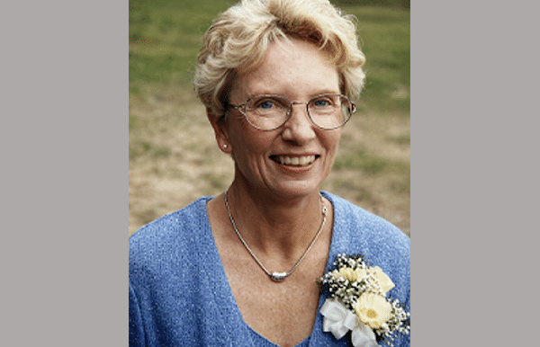 Close-up portrait of a woman with short blond hair, glasses and a blue dress, smiling in front of a grassy background.