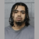 Man with medium-length locs and facial hair appears in a neutral booking photograph wearing a gray top.