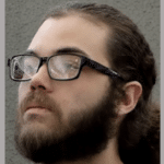 Bearded person in glasses gazes slightly upward against a textured gray background.