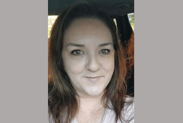 Portrait-style selfie of a brown-haired woman inside a vehicle, wearing a light-colored top and giving a subtle smile.