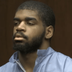 Head-and-shoulders image of a bearded man in a blue jail-issued top standing against a dark wood courtroom background.