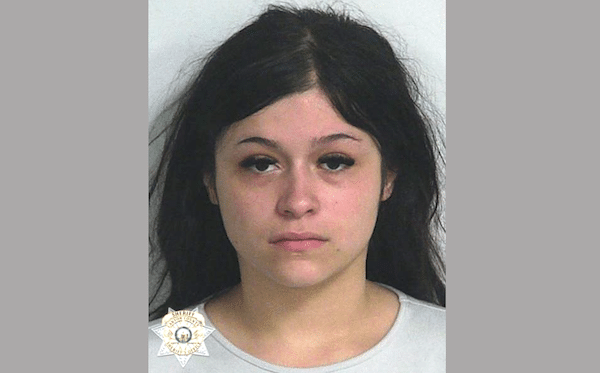 Close-up booking photograph of a woman with long black hair wearing a light-colored top against a pale backdrop.
